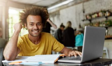 Happy positive African American college student with cheerful cute smile using wireless internet connection on laptop computer at coffee shop while looking for information online for research project