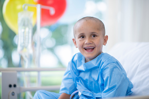 A young Ethnic boy is laying in a hospital bed. He has cancer, and his head is shaved. He is smiling happily.