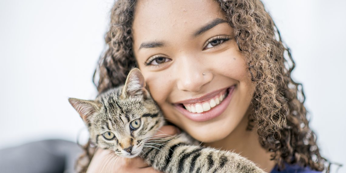 A teenage girl of African descent is indoors in her living room. She is holding up her newly-adopted cat, and smiling at the camera.