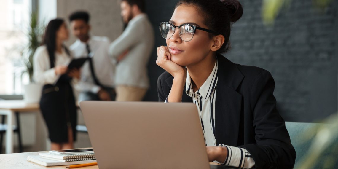 Photo of dreaming young african business woman sitting indoors using laptop computer. Looking aside.