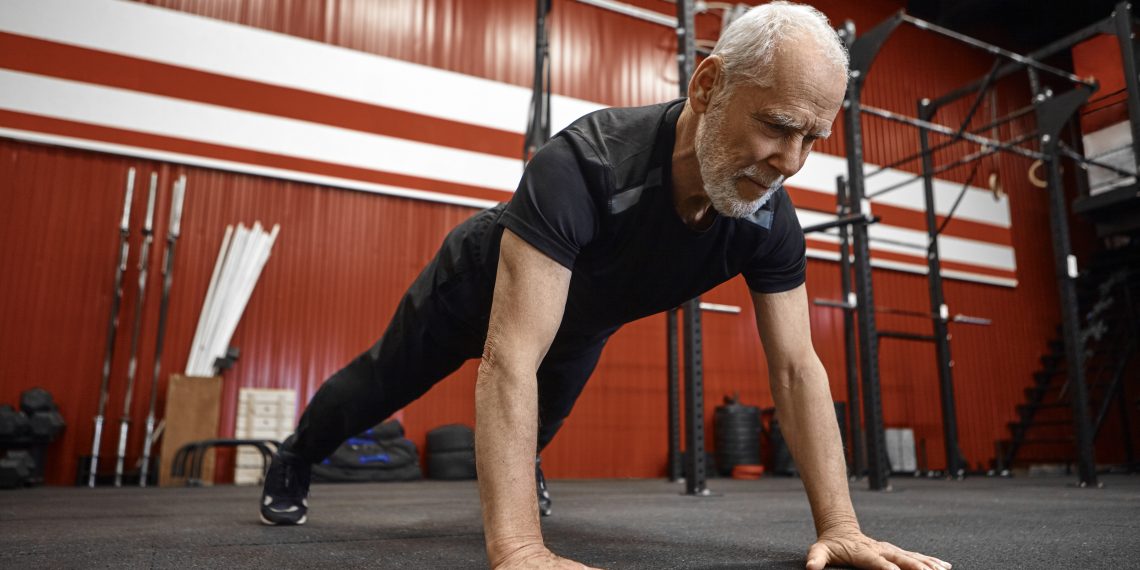 Healthcare, age, retirement and rehabilitation concept. Muscular fit seventy year old unshaven man in sportswear doing plank in gym. Senior male planking during morning workout at fitness center