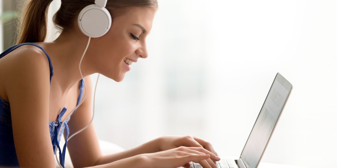 Side view portrait of smiling young woman in headphones typing on laptop while taking online course or personal training for self-education, qualification increase. Happy lady studying in Internet
