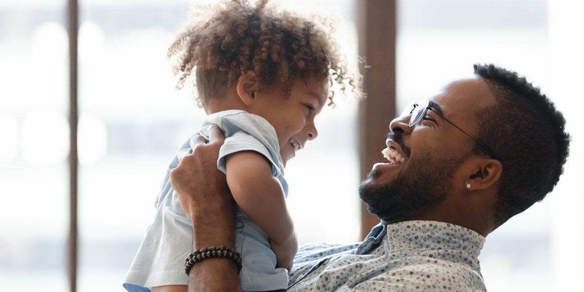 Happy daddy holding kid in arms, hugging toddler boy. African American dad enjoying being father, spending time and playing with cute preschooler son, talking to child, laughing. Parent concept