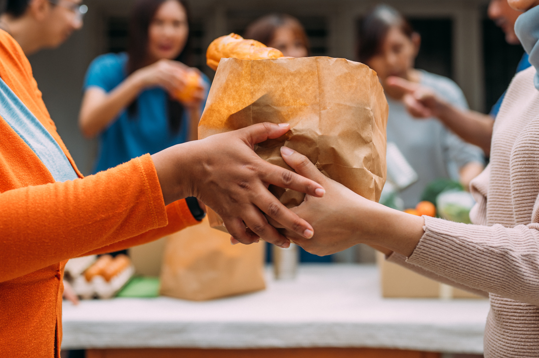 An unrecognizable woman hands a bag of donated food to someone during a community food drive.  An Asian female volunteer and her colleagues distributing grocery food at community food bank