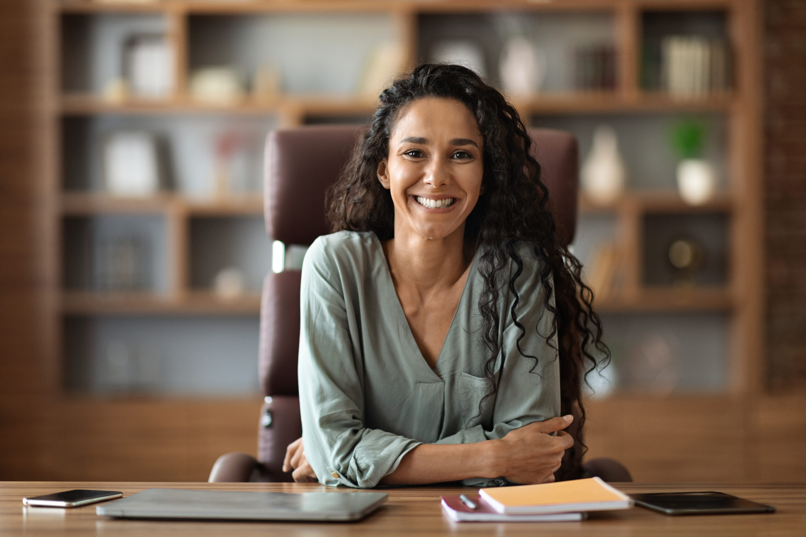 Happy attractive long-haired brunette young woman in casual outfit posing at office, female employee sitting at workdesk with gadgets and notepads on, cheerfully smiling at camera, copy space