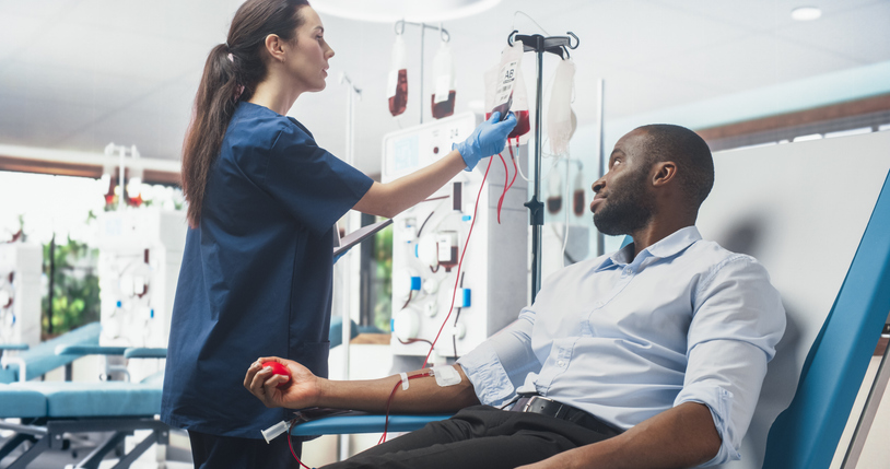 African Businessman Donating Blood For People In Need In Bright Hospital. Female Nurse With Tablet Computer Coming In To Check Progress And Well-Being Of Donor. Donation For Patients Battling Cancer.