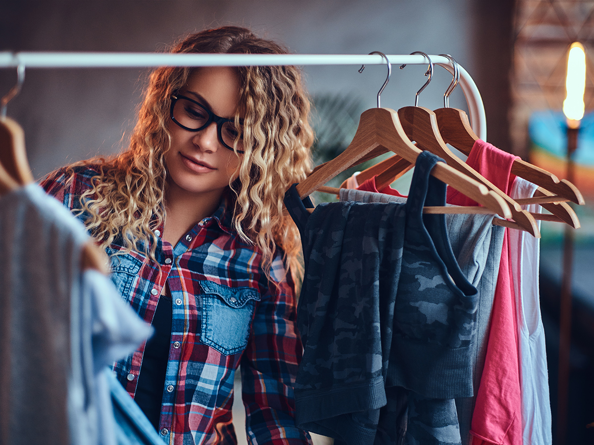 Positive blonde female in black eyeglasses chooses fashionable clothes on the coat rack.