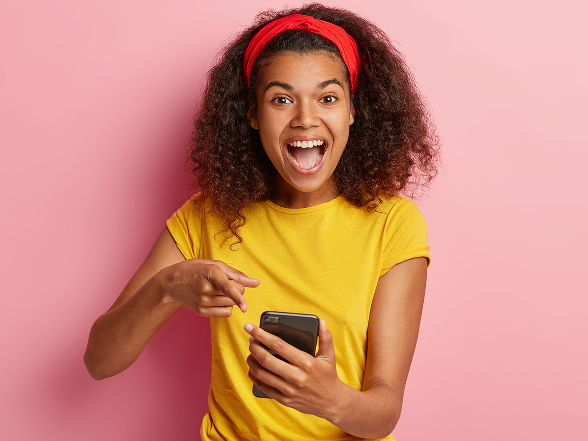 Joyful beautiful curly woman points at smartphone device with fast internet connection, views photos in social networks, wears yellow clothes, stands against rosy background. Selective focus.