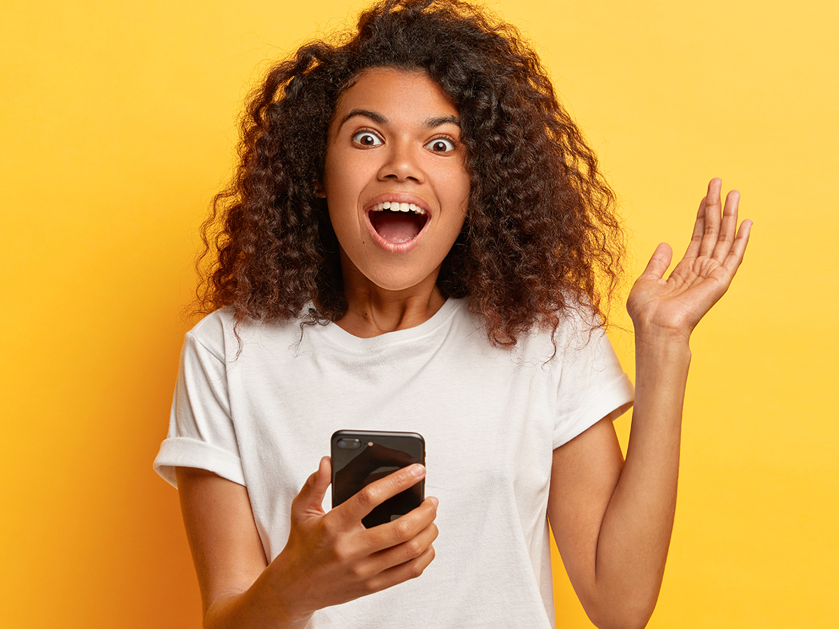Energized emotional Afro woman laughs as being entertained and amused, holds modern cellphone, raises arm, dressed in white t shirt and jeans, keeps breath from amazement, isolated on yellow wall